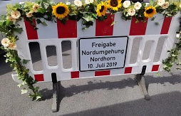 Auf einer mit Blumen geschmückten Sperrbake ist ein Schild angebracht mit folgender Aufschrift: Freigabe Nordumgehung Nordhorn, 10. Juli 2019.